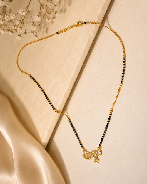A delicate gold chain necklace with small black beads and a butterfly-shaped pendant, styled on a cream-colored surface with satin fabric and dried flowers.
